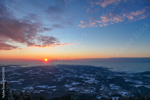 Fototapeta Naklejka Na Ścianę i Meble -  A sunrise seen from snow-capped peak of Schoeckl in Austrian Alps. The sky is bursting with orange and pink. Distant view on Graz, shrouded with fog. Winter wonderland. Day break. Calmness