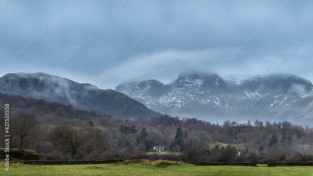 Epic dramatic landscape image of view from Elterwater across towards Langdale Pikes mountain range on foggy Winter morning
