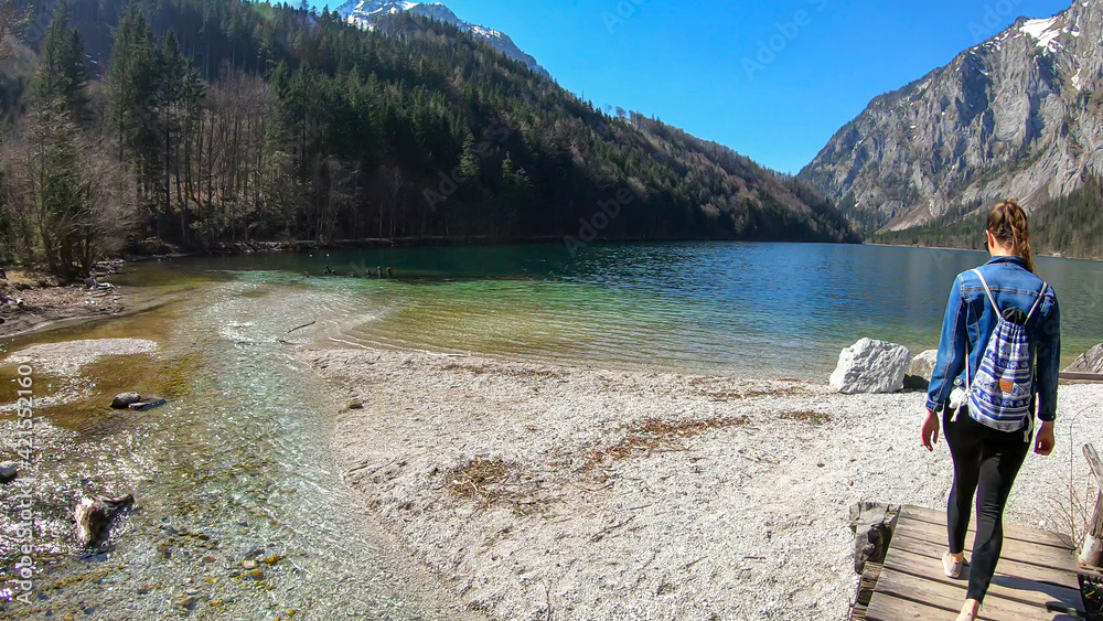 Fototapeta premium A woman walking along the shore of Leopoldsteiner lake in Austria. The lake is surrounded by high Alps. The shallow water is crystal clear, spring water has a calm surface. Early spring. Serenity