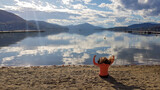 A woman in orange hoodie sitting at the sandy beach by the Woerthersee in Austria. The calm lake's surface is reflecting the surrounding mountains and clouds. Alpine landscape. The woman is meditating