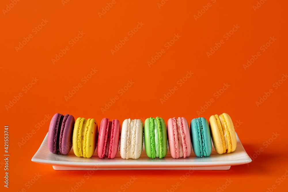 Multicolored macaroons in a row on a white rectangular plate against bright red background. Mockup of greeting card with colorful macaron cakes. Delicious french almond meringue cookies. Copy space.