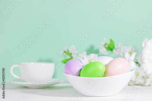 Easter eggs  pink  green and lilac  in a white plate  next to flowers with a cup of tea or coffee  on a green blue background. Pastel shades. Hard shadows. Easter breakfast.