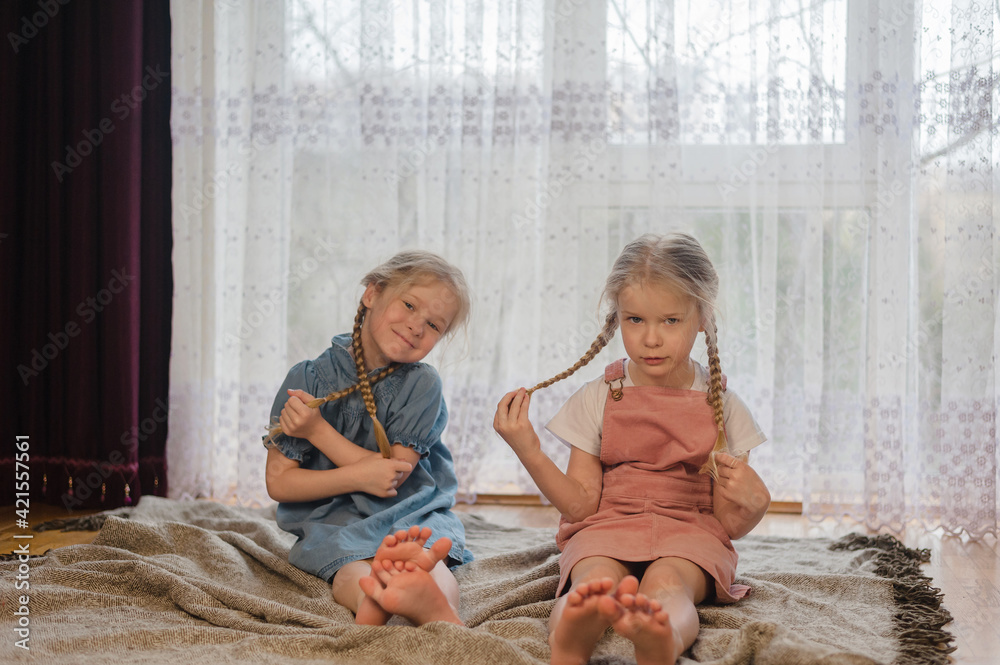 two little sister girls sit at home on the floor and play with their pigtails