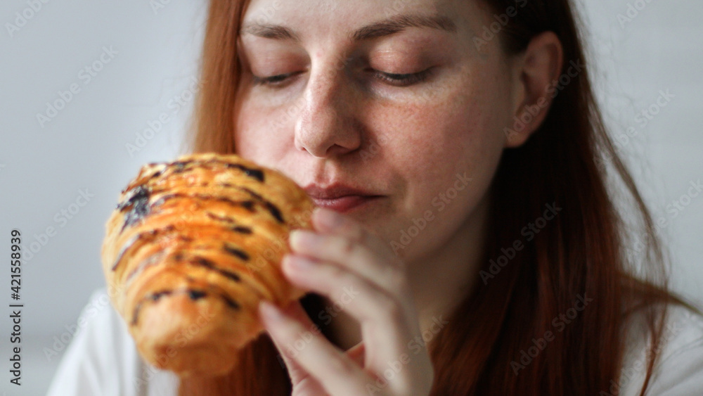 Happy young woman eats a croissant sitting on couch at home at morning
