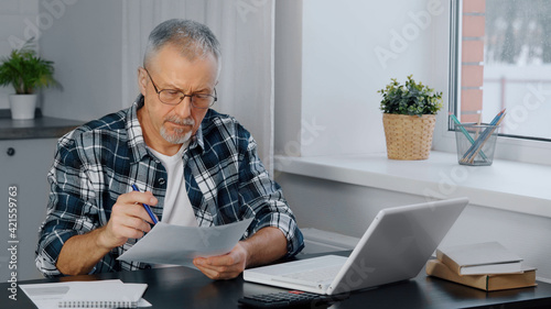 A thoughtful elderly man sits at his laptop, looking through documents.