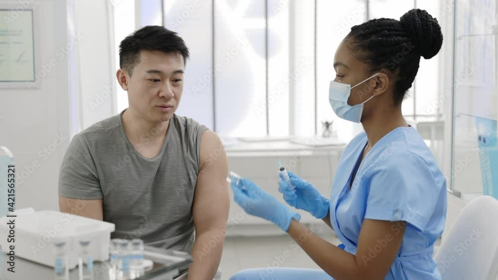 Young man communicating with doctor in protective mask and safety gloves while she filling syringe with vaccine in hospital while waiting for vaccination. Concept of prevention. Young man communicating with doctor in protective mask and safety gloves while she filling syringe with vaccine in hospital while waiting for vaccination. Concept of prevention.