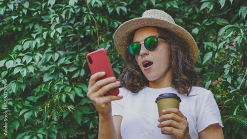 Excited young woman surprised by good unbelievable news, looking at smartphone screen feeling amazed, standing outdoors in green park, holding coffee cup to go. Unexpected win, shopping sale offer