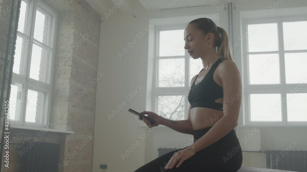 Low-angle medium shot with slowmo of attractive fit woman using smartphone while resting after hard indoor workout at gym looking at window Low-angle medium shot with slowmo of attractive fit woman using smartphone while resting after hard indoor workout at gym looking at window