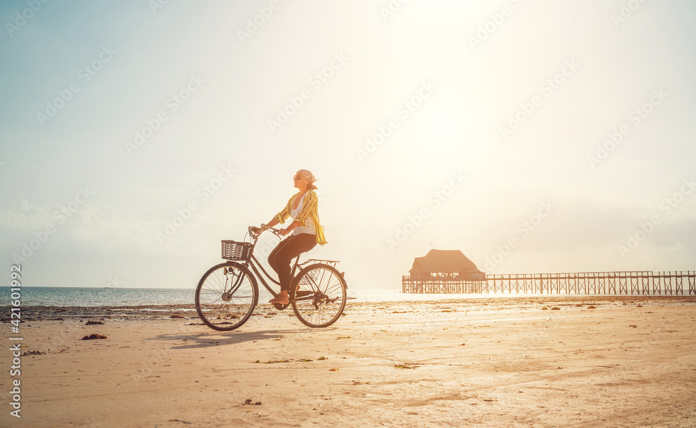 Fototapeta premium Young woman dressed light summer clothes riding old vintage bicycle with front basket on the lonely low tide ocean white sand coast on Kiwengwa beach on Zanzibar island, Tanzania.