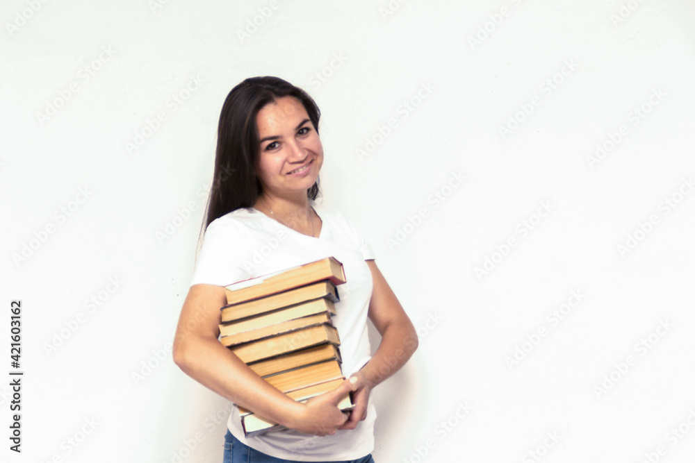 Young smiling student woman in white t-shirt holding textbooks and books in a pile on a white backdrop. College education concept. Positive emotions, facial expression Young smiling student woman in white t-shirt holding textbooks and books in a pile on a white backdrop. College education concept. Positive emotions, facial expression