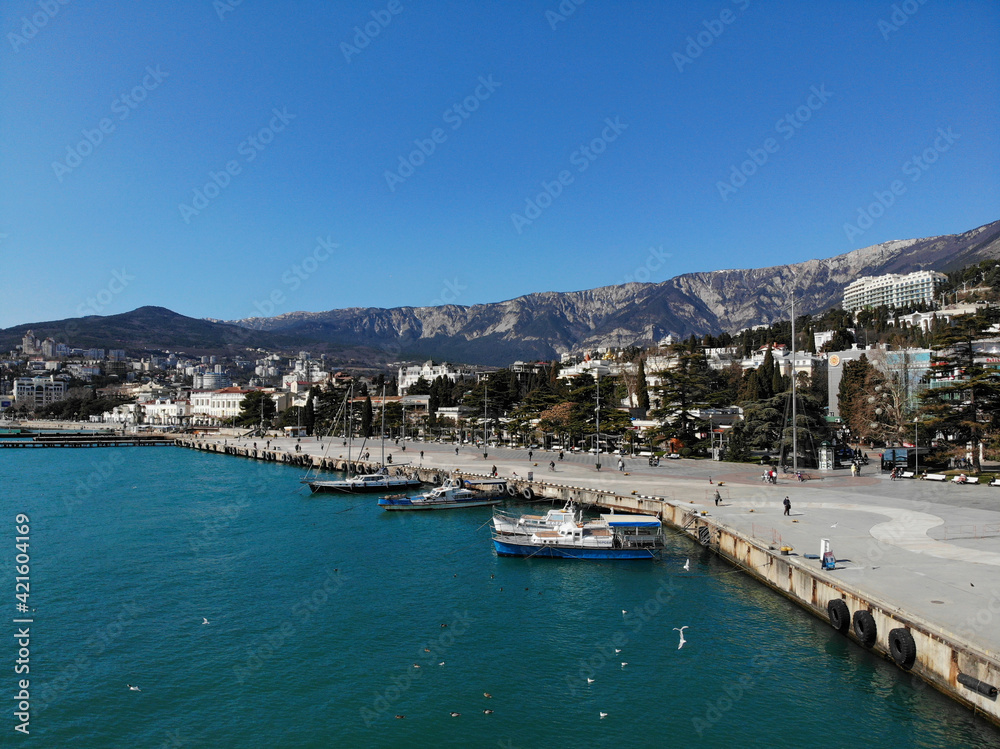 Aerial view of the panorama of dock with moored boats or yachts, resort city Yalta and the Crimean mountains at sunny spring day from sea, from above