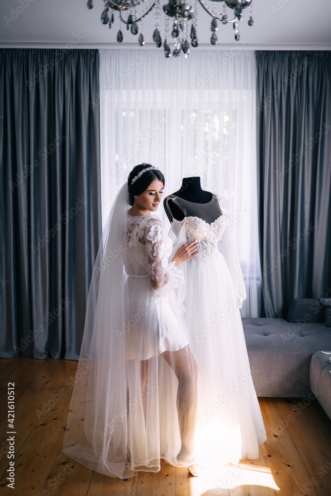 A beautiful, dark-haired bride in lingerie, in a robe hugs a hanging white lace dress.