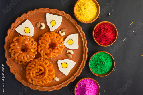 Colorful holi powder or gulal in earthen bowl along with traditional Indian sweet food imarti, sandesh, cashew and almonds in plate on black background. photo