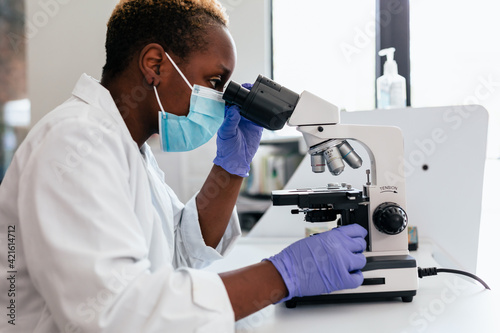 Black female scientist in mask in laboratory looking through microscope
