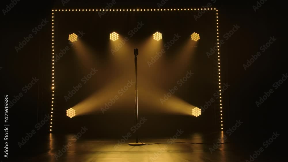 A shiny old retro microphone stands on an empty stage in the dark. Music equipment in a smoky studio lit with bright stage spotlights and soft yellow lights. Slow motion.