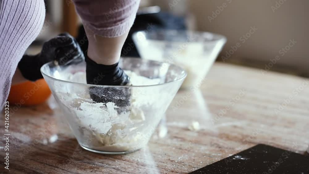 Una mujer en la cocina amasando una masa de pizza dentro de un bol con harina mientras usa guantes negros.