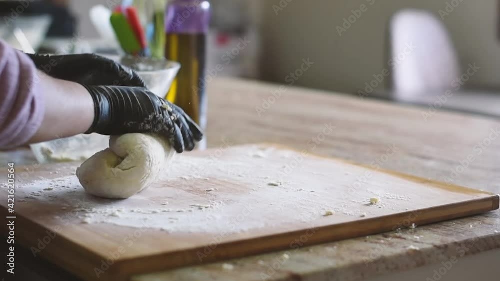 Una mujer en la cocina amasando una masa de pizza en una tabla de madera con harina mientras usa guantes negros.