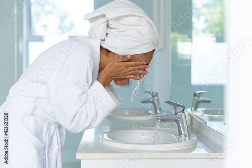 Mixed race woman wearing bathrobe and washing her face in bathroom