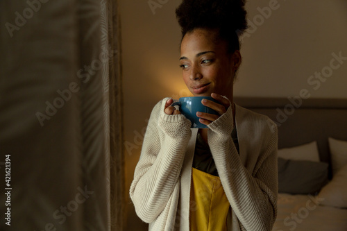 Mixed race woman standing by a window and drinking cup of coffee at home