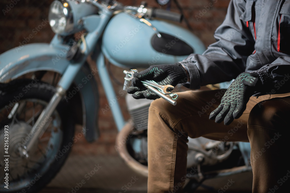 A mechanic with a wrench in hand is sitting on the broken motorbike background and is spreading his hand. Motorcycle repairing service concept. A mechanic with a wrench in hand is sitting on the broken motorbike background and is spreading his hand. Motorcycle repairing service concept.