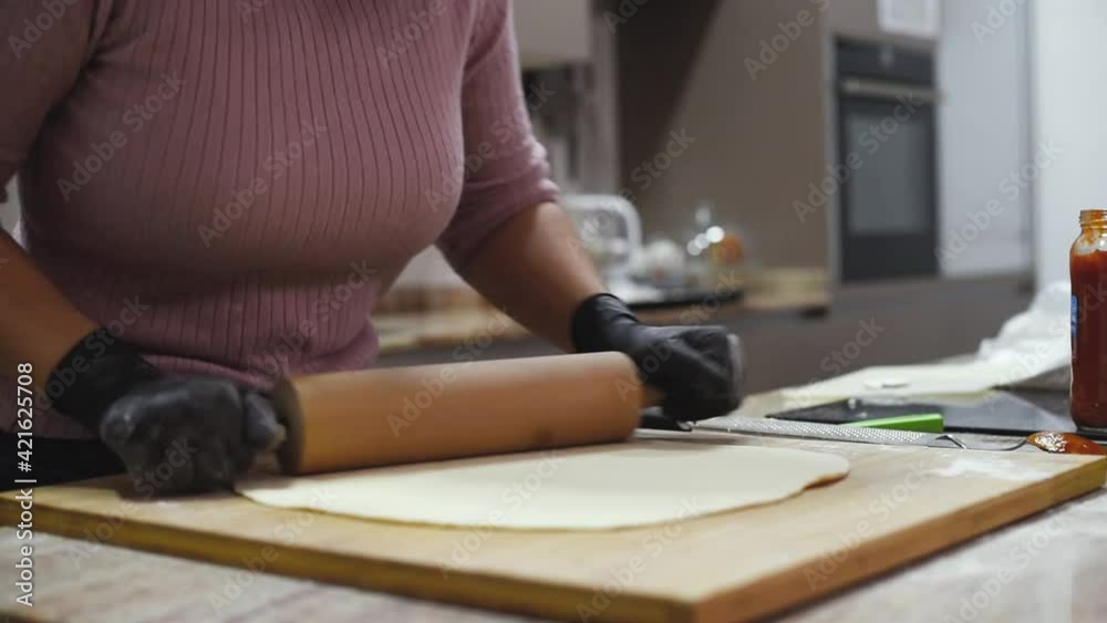 Una mujer con guantes negros en la cocina usando un rodillo de madera para amasar y estirar una masa de pizza en una tabla de madera. Concepto de hacer pizza casera y artesanal en casa