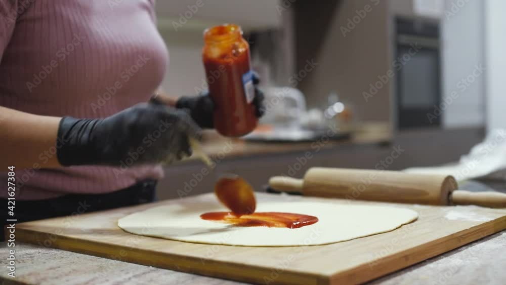Una mujer con guantes extendiendo salsa de tomate con una cuchara sobre una base de pizza casera. Bote de salsa de tomate.