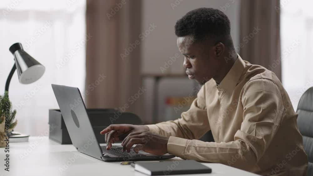 afro-american guy is working with laptop at home, young freelance worker is looking on display and typing afro-american guy is working with laptop at home, young freelance worker is looking on display and typing