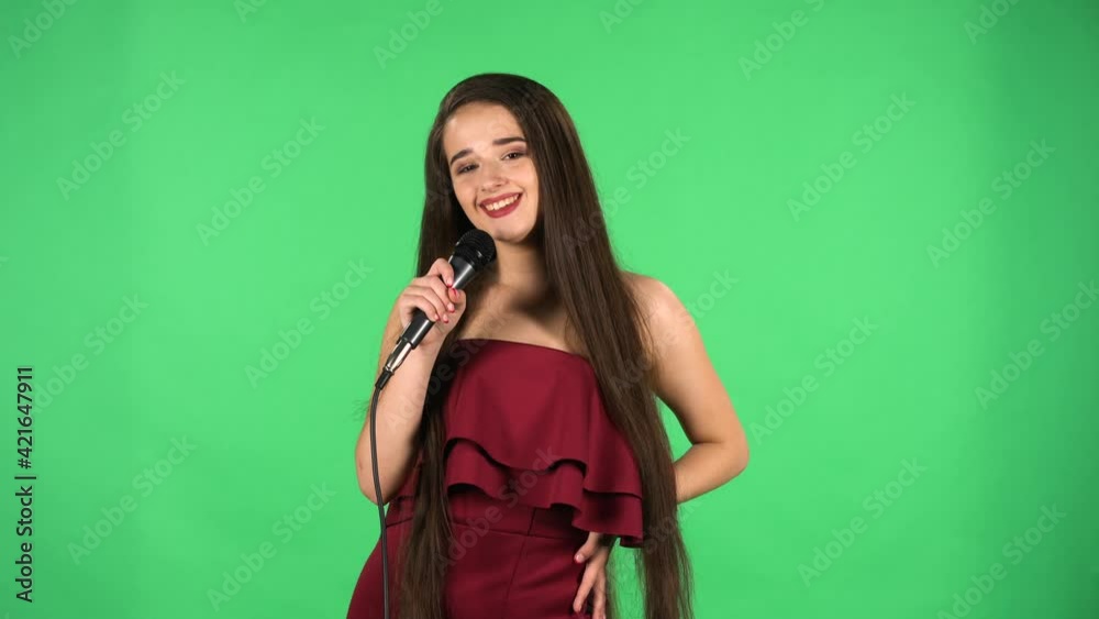 Portrait of beautiful young woman coquettishly looking at camera and sings song into microphone. Model with long hair in red dress posing on green screen in the studio. Slow motion. Close up. Portrait of beautiful young woman coquettishly looking at camera and sings song into microphone. Model with long hair in red dress posing on green screen in the studio. Slow motion. Close up.