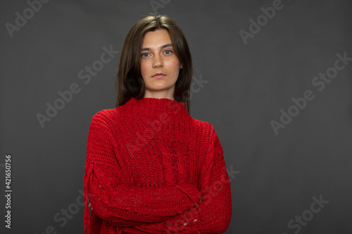pretty long-haired brunette girl posing with crossed arms in red sweater looking at camera on gray studio backdrop copy space. Calmness, confidence concept. tacit consent, approval, appeal in her eyes