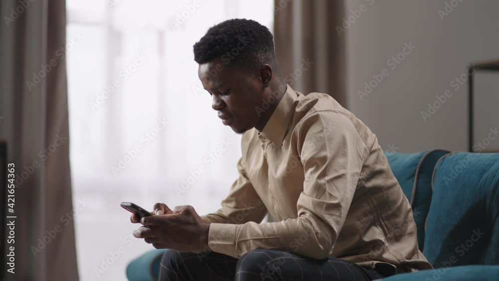young man is sitting in apartment and using smartphone, afro-american guy is surfing internet by mobile phone at home, resting alone at weekend