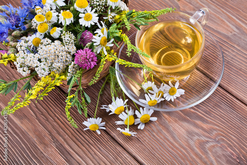 Herbal tea with the chamomile flowers in a glass cup and fresh wild flowers in a basket on a wooden background.
