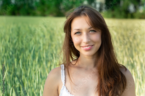 Attractive smiling girl in summer dress with open shoulders and magnificent dark hair looking at camera on blurred green wheat field background.