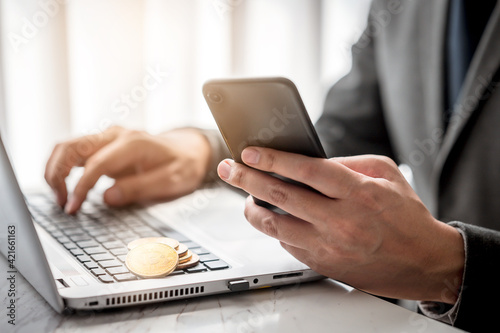 Close up shot of businessman hand using smartphone and laptop with golden bitcoin on keyboard