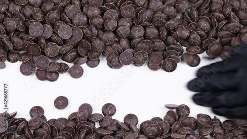 chefs hand in black glove shovels Dark chocolate couverture callets on white table. Chef or chocolatier preparing to melt chocolate. Cocoa processing, Chocolate production concept.