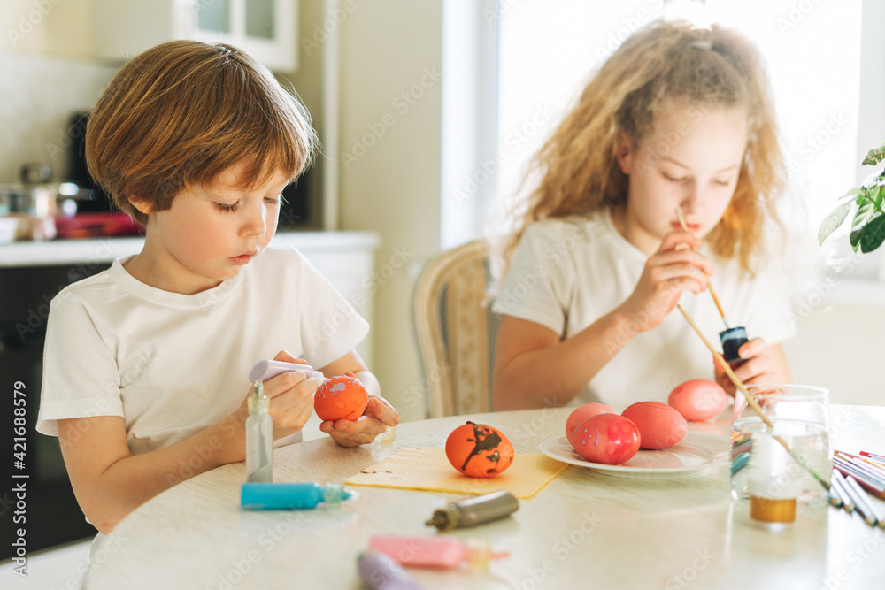 Two siblings brother and sister toddler boy tween girl painting easter eggs on kitchen at home on spring sunny day Two siblings brother and sister toddler boy tween girl painting easter eggs on kitchen at home on spring sunny day