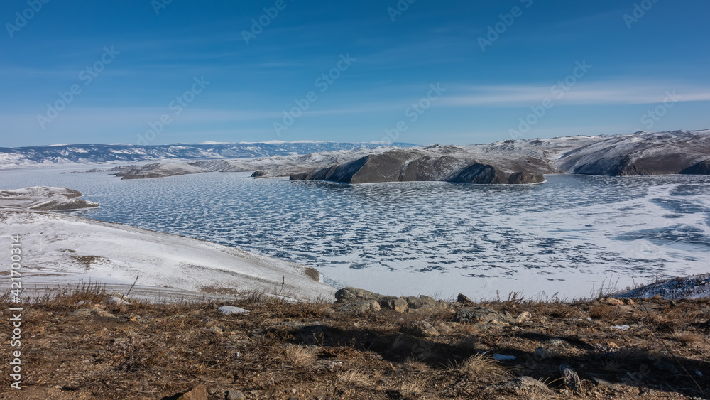 The frozen Lake Baikal is covered with lace-like snow. In the foreground is the earth, dry grass. In the distance, snow-capped mountains. Blue sky. Winter day. The frozen Lake Baikal is covered with lace-like snow. In the foreground is the earth, dry grass. In the distance, snow-capped mountains. Blue sky. Winter day.
