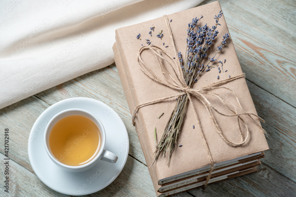Neat, beautiful stack of books with craft paper covers, wrapped in rope and decorated with lavender flowers, and a cup of tea on a wooden table Neat, beautiful stack of books with craft paper covers, wrapped in rope and decorated with lavender flowers, and a cup of tea on a wooden table