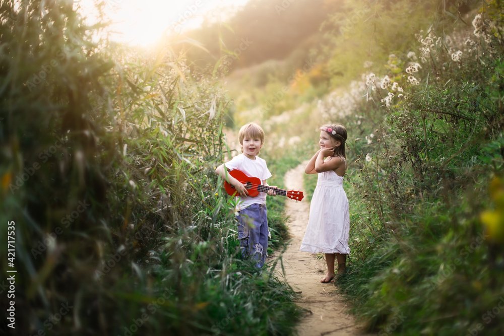 Fototapeta premium Cute romantic children a couple, a boy plays the guitar and sings, a girl in a dress is dancing. Children in a summer park barefoot on the grass. happy carefree childhood and romance