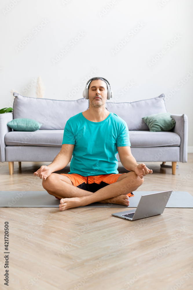 Man meditates at home - using from the internet to monitor the workout. He listens to relaxing meditation music through headphones.