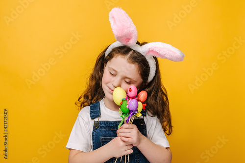 Portrait of dreamy carefree pretty charming preschool girl kid with closed eyes holds small colored eggs on sticks, wears pink bunny fluffy ears, isolated on yellow background. Easter holiday concept