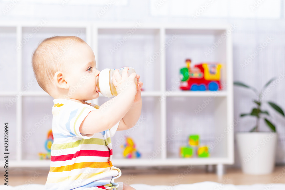 Cute caucasian baby boy drinking from bottle fruit tea with camomile. Sitting on white carpet in striped bodysuit. Different children's toys in the background. Side view Cute caucasian baby boy drinking from bottle fruit tea with camomile. Sitting on white carpet in striped bodysuit. Different children's toys in the background. Side view