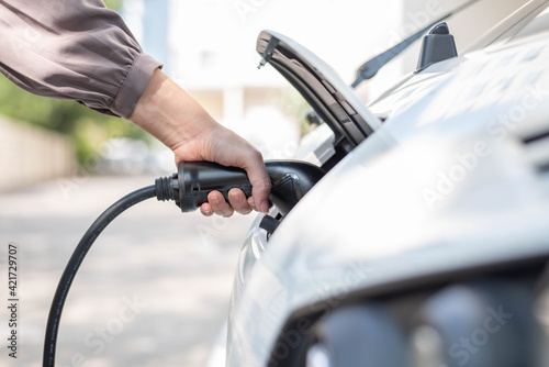 Close up, side view shot, selective focus of a woman's hand grabbing, plugging, holding the EV charger plug handle in front of an electric car. Sustainable energy concept