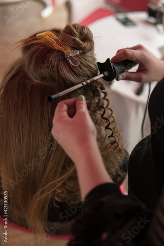 Close up shot of female stylist making curly hairstyle of female model with curling iron device.