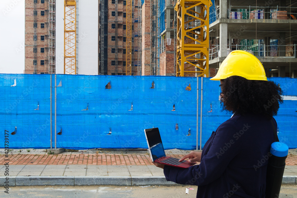 african-american woman construction manager in yellow hardhat set out to check the construction of the flats. The woman is holding a laptop computer in her hands