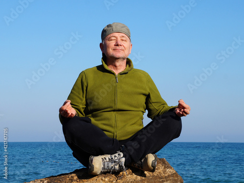 middle aged man sits in lotus position on a large rock by the sea and meditates in sunlight photo