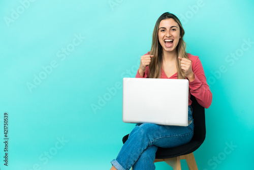 Young caucasian woman sitting on a chair with her pc isolated on blue background celebrating a victory in winner position © luismolinero