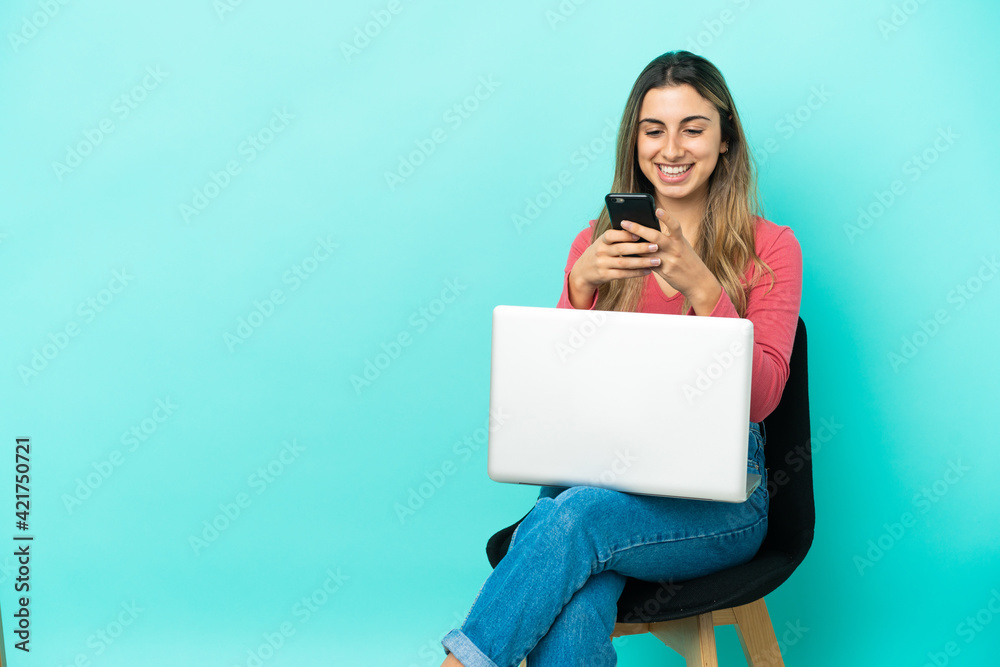 Young caucasian woman sitting on a chair with her pc isolated on blue background sending a message with the mobile Young caucasian woman sitting on a chair with her pc isolated on blue background sending a message with the mobile