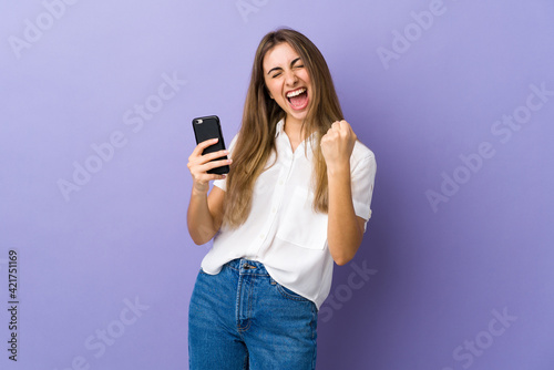 Young woman over isolated purple background with phone in victory position