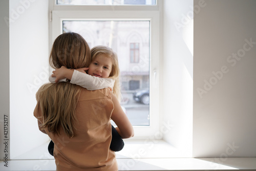 Beautiful young woman and her charming little daughter are hugging and smiling at home