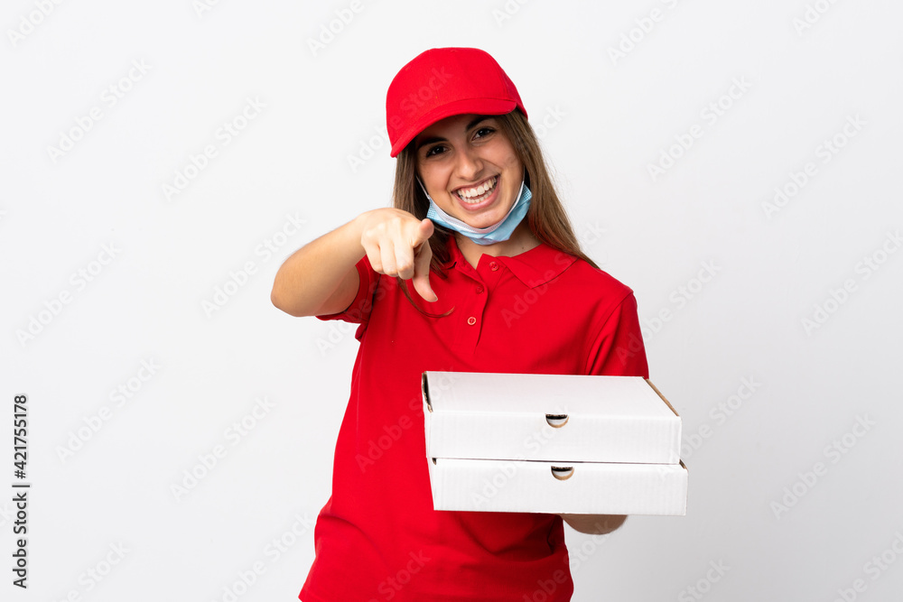 Pizza delivery woman holding a pizza and protecting from the coronavirus with a mask over isolated white wall points finger at you with a confident expression Pizza delivery woman holding a pizza and protecting from the coronavirus with a mask over isolated white wall points finger at you with a confident expression